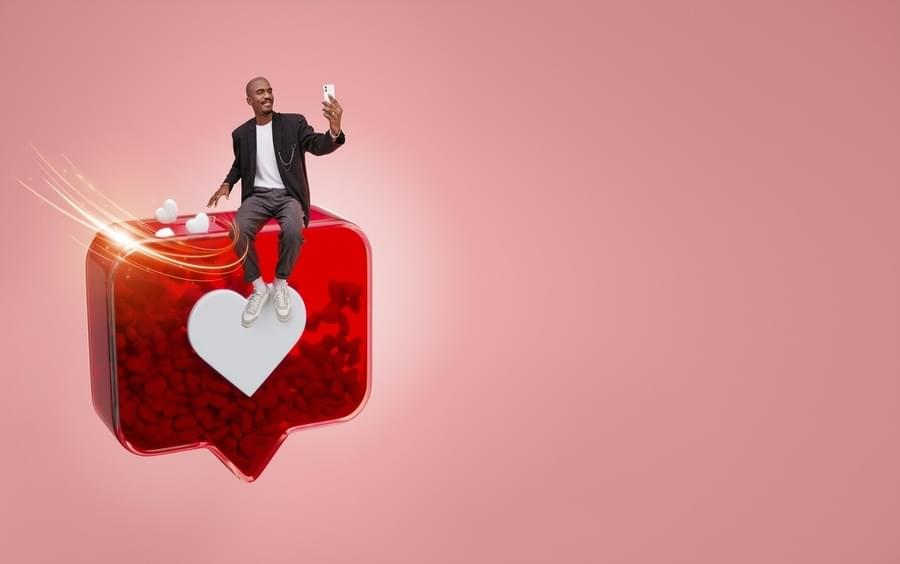 Man sitting on a large 3D Instagram-like icon with a white heart symbol, taking a selfie against a pink background with floating heart elements